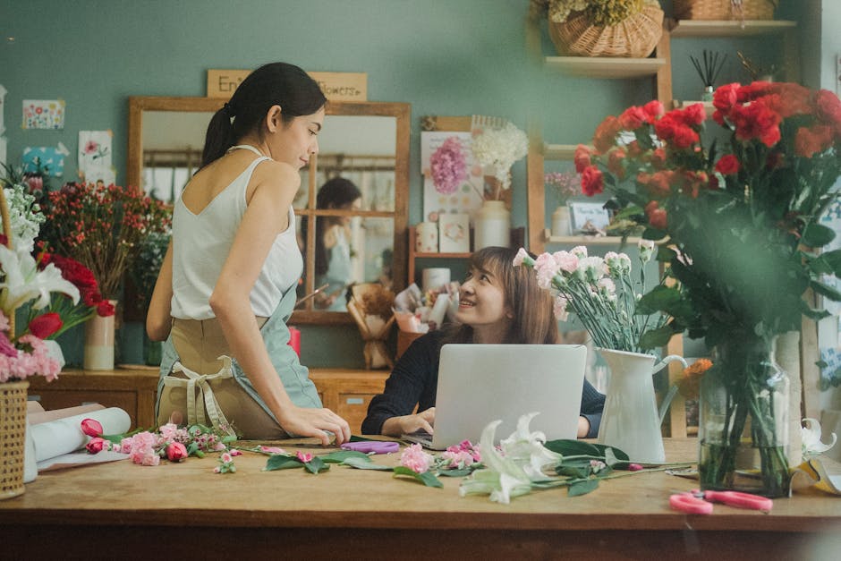 Inside a flower shop with soft green walls, two women are engaged in a floral arrangement. One woman, standing, has dark hair tied back and is wearing a white sleeveless top and a beige apron. She is