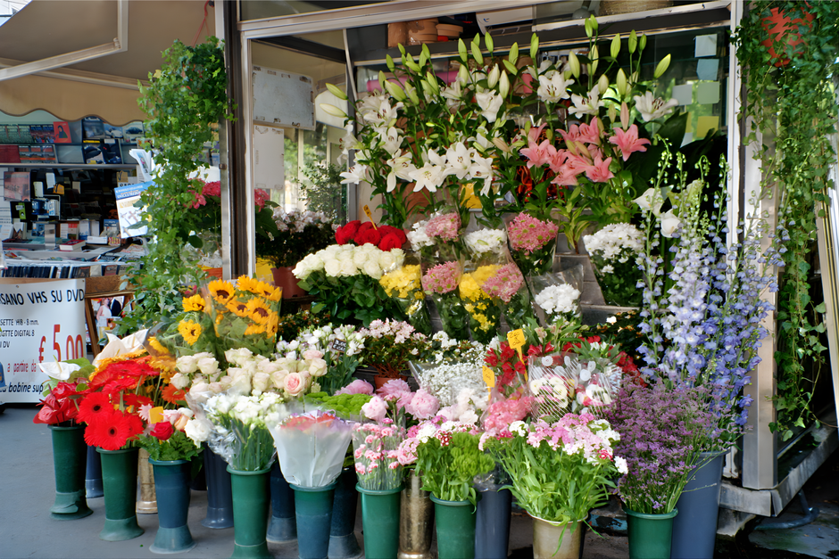 A vibrant display of fresh flowers outside a florist shop, including white lilies with large open petals and green stems at the top, pink lilies with smaller blossoms, and various other colorful bloom