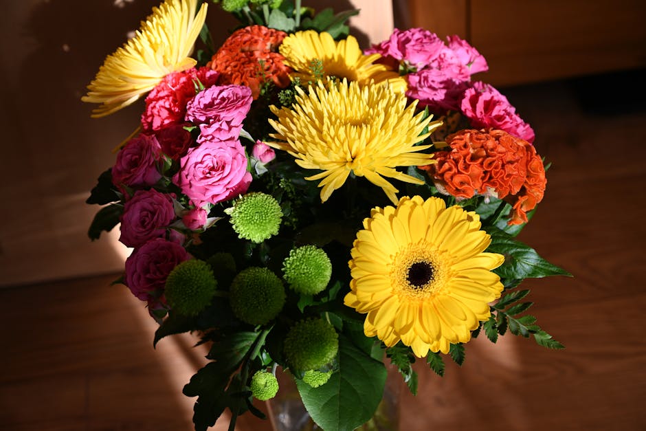 A vibrant floral bouquet arranged in a clear glass vase, featuring large yellow gerbera daisies with dark centers, bright pink roses in full bloom, orange carnations, and small green chrysanthemum blo