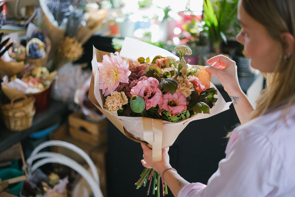 A woman with long, light brown hair partially tied back, dressed in a light-colored top, is holding a bouquet of fresh flowers wrapped in beige and white paper with a cream-colored ribbon. The bouquet