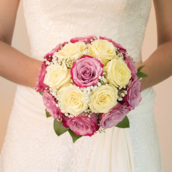 The camera lingers on a bride's hands, gently supporting a round, sumptuous bouquet that blossoms with wedding-day promise on the edge of Beckton Park. Roses, the main protagonists, unfold in dense clusters-most displaying a soft, buttery yellow tone, others glowing in vibrant fuchsia or tinged with delicate mauve, their gradients lending rich texture and depth. A constellation of tiny white Gypsophila, or baby's breath, threads through the roses, evoking the delicate springtime air off Royal Docks, while a few bold, verdant leaves emerge at the periphery. The bride's arms create a graceful arch around the bouquet, the understated sophistication of her ivory gown and its intricate lace motifs subtly visible. The fabric falls softly, catching the light and echoing the bouquet's gentle curves. The atmosphere is serene-the bright, even lighting accentuates both freshness and complexity, drawing the eye to every bloom and lace detail. Faintly blurred, the background hints at a quiet morning in Beckton, turning all focus inward to the joyful centrepiece. Crafted for storytelling on wedding planning or floral design, this photograph captures a fleeting but unforgettable moment-a testament to the artistry and care found in Beckton's weddings.