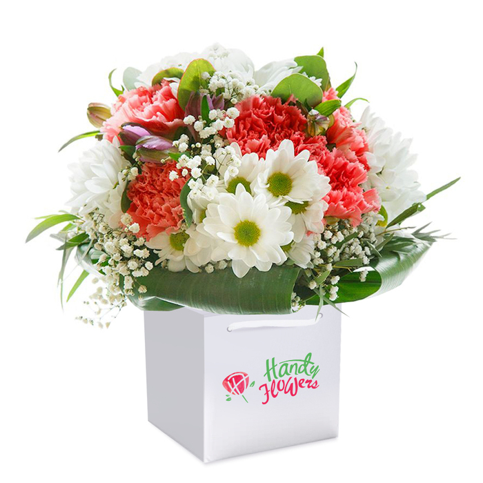 Resting upright within a bright, square white gift bag, this radiant bouquet is a celebration of texture, colour, and freshness prepared by Beckton Flowers for families and friends throughout the Beckton area. Soft coral and peach carnations, each petal vividly ruffled and full of lively movement, anchor the bouquet with a sense of richness and warmth. Among them, rich red carnations add a passionate, festive energy, while clusters of elegant white daisies with vibrant yellow-green centers offer a fresh, cheerful contrast that calls to mind the open, sunlit spaces of Beckton's parks. Airy sprigs of delicate baby's breath are interspersed lightly, lending the arrangement a gentle, cloud-like quality and a light fragrance evocative of early mornings on the nearby Greenway. A touch of mystery arrives in the form of unopened, purple-tinged buds emerging from one side, adding intrigue and suggesting that more beauty is yet to blossom. All blooms are gracefully encircled by shiny, deep green leaves, whose smooth surfaces and broad shapes frame the composition and provide a natural, grounded elegance. The white gift bag is refined and functional, with strong white cord handles positioned for effortless local delivery or personal gifting, whether it's arriving on a doorstep along Winsor Terrace or brightening a table for a family gathering. Every flower is artfully positioned, and the spirited arrangement is intended to inspire connection and joy, making it perfect for special celebrations, milestones, or just to brighten a day in Beckton.