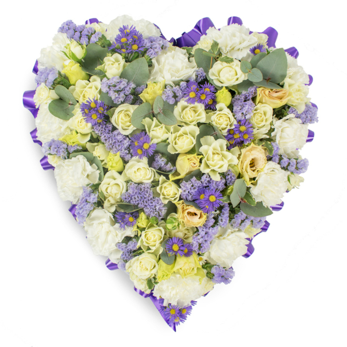A meticulously crafted heart of flowers, photographed against pristine white to let the arrangement's gentle hues speak plainly: creamy and pale yellow roses crowded together to form a sumptuous centre, each bloom showing varying stages from tight bud to soft open cup, giving the piece a layered, intimate texture. Nestled among the roses, voluminous white carnations add a traditional, consoling fullness, their ruffled petals creating cloud-like pockets of light. Interspersed throughout are clusters of lavender statice, their tiny, papery florets lending a misted purple veil, and vivid small purple asters with bright yellow discs that act as cheerful but restrained accents against the softer tones. Silvery-blue, rounded eucalyptus leaves appear at intervals, breaking the floral mass with cool colour and a hint of fresh, herbaceous fragrance; their shape adds balance and a contemporary touch. The heart's edge is finished with a continuous, deep purple satin ribbon folded into neat loops, providing a refined border that clearly defines the shape on the white backdrop. The lighting is soft and even, accentuating petal texture and the gentle contrast between cream and lavender.
This arrangement reads as a peaceful statement of sympathy-appropriate for funerals, memorial services or placing near a gravestone in the Beckton or Gallions Reach area-and is intended to comfort mourners with its quiet palette and considered craftsmanship, symbolising devoted remembrance and heartfelt condolence.