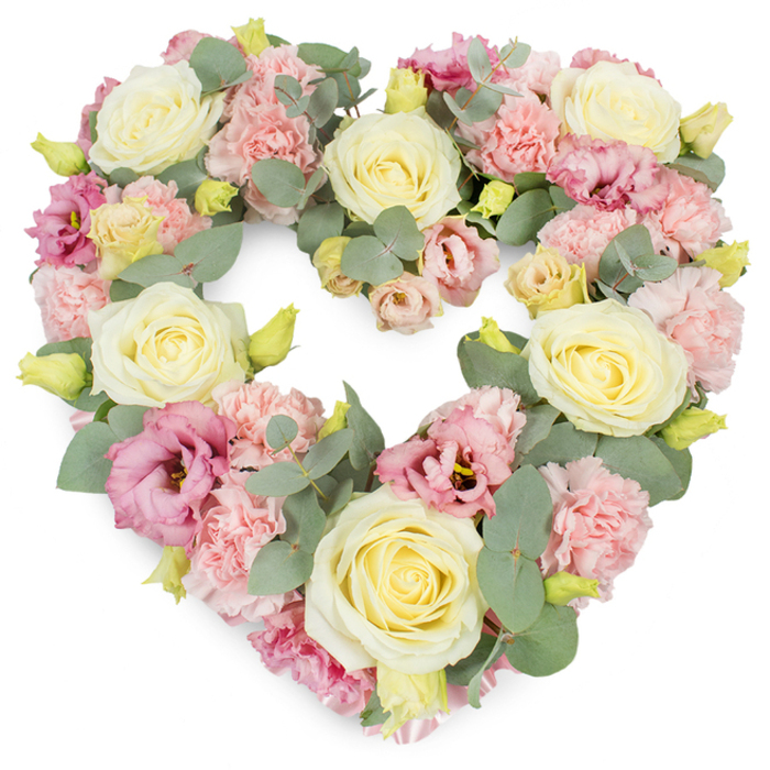 A meticulously crafted heart-shaped wreath sits against a pristine white backdrop, its pastel tones and lush texture drawing the eye into a gentle, reflective space. The heart is fully formed and symmetrical, built from a base of large, creamy ivory and pale lemon roses, each bloom with tightly furled inner petals that slowly open into a soft, rounded cup. These roses provide structure and a sense of calm, anchoring the design with their understated elegance. Between them, countless ruffled pastel pink carnations spill into the gaps, their frilly edges overlapping to create a plush, almost fabric-like surface that feels comforting and warm. Deeper pink to mauve lisianthus are scattered thoughtfully throughout the wreath, some opening wide, others still partly curled, adding variety in both shape and tone. Cool, silvery-green eucalyptus stems twist gently around the flowers, their round leaves and slightly dusty finish offering a soothing contrast to the smooth rose petals. Small yellowish-green buds peek out here and there, contributing discreet touches of brightness and conveying the freshness of a design recently prepared. The flowers are placed so closely together that the wreath appears dense and rich, echoing the depth of feeling behind such a tribute. Imagined in the hands of an experienced Beckton florist, this heart could be destined for a funeral service at City of London Cemetery or a memorial gathering at a local chapel, offering a dignified, pastel expression of love, sorrow, and remembrance. Its soft pinks, creams, and greens work together to convey compassion and the quiet reassurance of enduring affection.