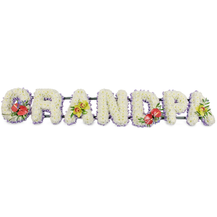 Set against a bright, uncluttered white backdrop, this personalised funeral flower arrangement spells the word GRANDPA in prominent block capitals, each letter formed from a dense carpet of small white chrysanthemums. The blooms are placed so closely together that no gaps show, creating a smooth, velvety texture that reads as pure and serene, a fitting symbol of peace and remembrance. Around the bottom edge of each letter runs a slim band of light lavender ribbon, just visible beneath the white flowers and providing a gently tinted border that lifts the design from the background without distracting from its calm simplicity. All seven letters are connected along a discreet dark green frame, giving the tribute enough strength to rest atop a coffin, stand at the front of a chapel, or be displayed at a graveside service in Beckton. Carefully chosen accent sprays bring colour and meaning: the G and the D carry bold groupings of deep red anthuriums, their polished, heart-shaped petals and central yellow spadices surrounded by glossy green foliage and a few scattered purple filler blooms that echo the ribbon trim. The first A and the last A are softened with clusters of yellow-green orchids, their delicate, arching shapes and subtle markings offering a sense of gentle grace. The interplay of white, red, purple, green, and orchid tones ensures the tribute feels rich yet respectful. It is the kind of bespoke Grandpa floral lettering a family in Beckton might order to honour a grandfather's life, combining traditional sympathy flowers with a personalised message of enduring love.