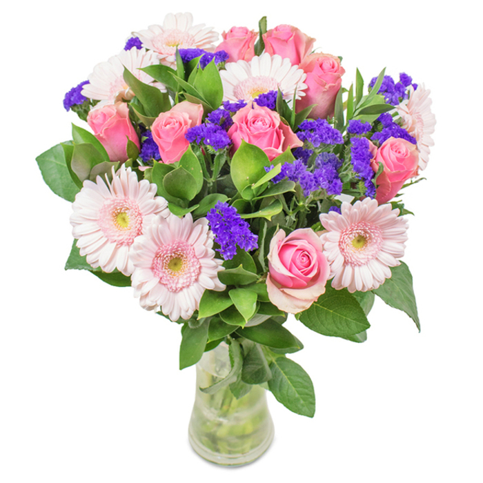 A visually striking Beckton bouquet stands poised in a pristine clear glass vase, set against a crisp white background that enhances its rich colour story. At its heart, approximately 7-9 soft and medium pink roses display a mix of tightly coiled buds and delicately unfurling petals, each capturing the classic charm of a traditional English rose. Interwoven are 5-7 pale blush gerbera daisies, large and radiant with gentle faces and lighter, nearly white centers-an echo of early blooms in Beckton's local parks. Between the main blooms, vivid clusters of deep purple statice introduce a playful, textural contrast, their intense colour drawing the eye and invigorating the arrangement. The base is woven with abundant deep green foliage, creating an impression of local meadows at their most lush and offering hints of fresh, dewy scent. The transparent vase reveals healthy green stems and clean water, suggesting freshness and local care with every detail. Whether delivered for a birthday, an anniversary, or simply to uplift a Beckton neighbour's home or office, this distinctive combination of roses, gerberas, and purple accents embodies celebration, joy, and meaningful connection.