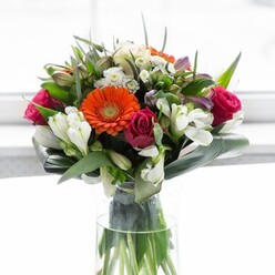 This exquisite bouquet, arranged by expert hands just a stone's throw from Beckton's bustling shopping centres, is a study in textured elegance and radiant colour. At the forefront, two dazzling orange gerberas ignite the composition, their fiery petals capturing attention like the sunset seen from Beckton Pier. Soft peach and creamy-beige gerberas encircle these, their gentle hues whispering calm and comfort, evocative of quiet mornings in neighborhood gardens. Interspersed among the gerberas, bold fuchsia roses wind their way through, introducing layers of lush romance and sophistication, while clusters of snowy alstroemeria and petite white spray roses break through the greenery, imparting an immaculate brightness that accentuates the arrangement's abundant feel. Small, light pink blossoms and sprigs of delicate white filler flowers nestle between larger blooms, deepening the overall texture and evoking the subtle complexity of a local wildflower meadow. A luxurious cascade of glossy, deep green foliage-broad paddle-shaped leaves elegantly arched and wrapped-cradles the bouquet, forming a natural collar that brings structure and tranquility to the lively colours above. Smaller pointed leaves and finely textured greenery fill every space, their fresh shades echoing the vibrancy of Beckton's open spaces. The tall, transparent glass vase anchors the softness of the bouquet, displaying the verdant stems beneath and heightening the sense of natural abundance. Illuminated by soft, diffused light, every bloom and leaf glows with a freshness that suggests careful curation and pride. Perfect for brightening a table on Winsor Terrace or marking a special moment with a neighbour, this arrangement communicates hope, gratitude, and the enduring joy of flowers delivered from the heart of Beckton.